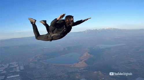 A skydiver dressed in a black jumpsuit descends against a clear blue sky, with distant mountains and a body of water framing the scene. The YouTube Originals logo is positioned at the bottom right.