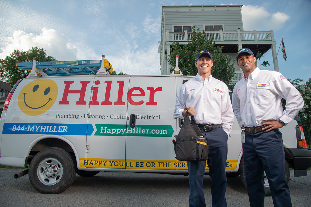 Two uniformed technicians stand in front of a Hiller Plumbing Company van parked outside a home. The van prominently features the Hiller logo, contact information, and signature yellow smiley face branding. One technician is holding a black tool bag, demonstrating readiness for service.