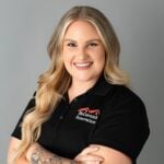 Alexa Nicholas, sporting long blonde hair and a confident smile, stands with arms crossed. She dons a black polo shirt showcasing the "McCormick Renovations" logo in front of a neutral gray background.