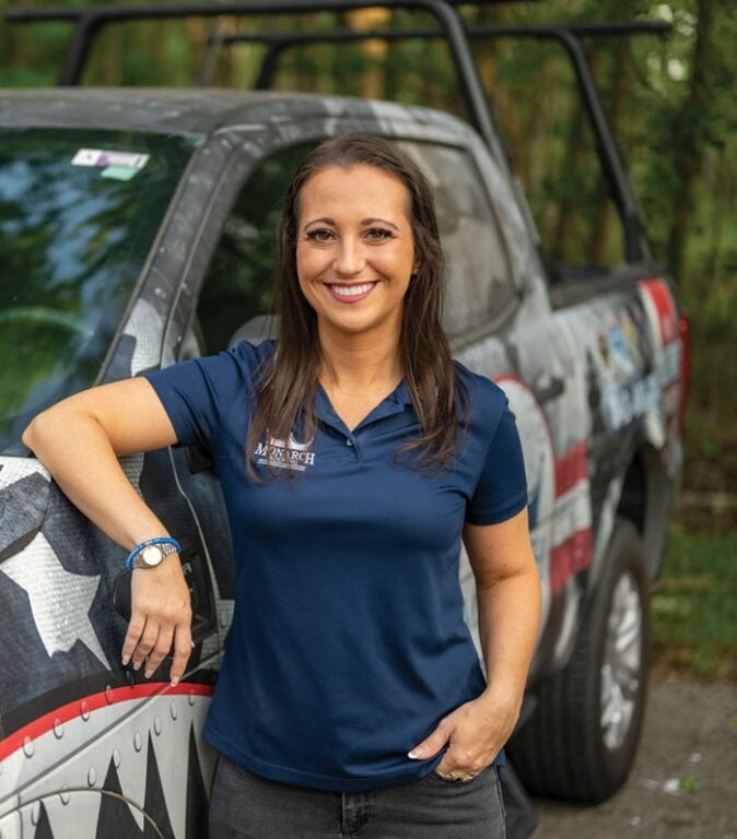 A woman, Alicia Tretault of Monarch Roofing, in a blue polo shirt stands smiling with one hand on a truck adorned with a star-spangled design, showcasing her roofing success story. With long brown hair flowing, she radiates confidence outdoors against a wooded backdrop—a living case study in marketing prowess.
