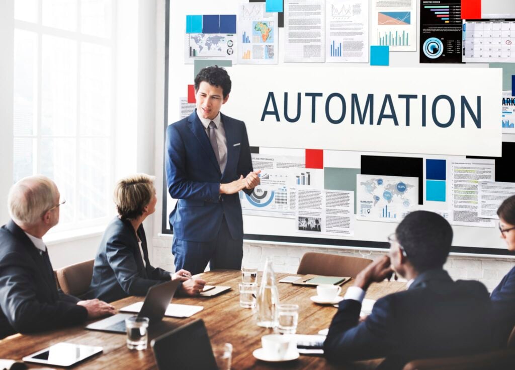 A professional in a business suit delivers a slideshow presentation on "Automation" to colleagues gathered around a conference table. The backdrop features charts and graphs illustrating how automation can enhance profitability.