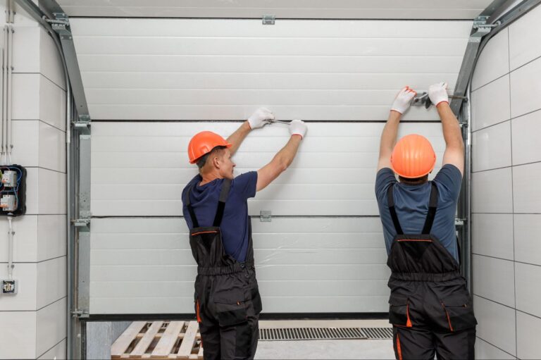 Two workers in orange helmets and dark overalls install a white garage door, using tools inside a garage for Hook Agency clients.