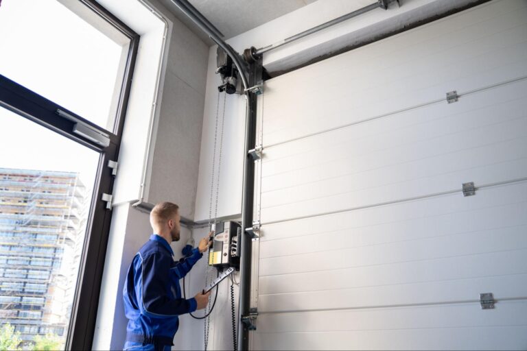 Technician in blue overalls inspects and adjusts the control panel of a large white industrial garage door, construction site seen outside.