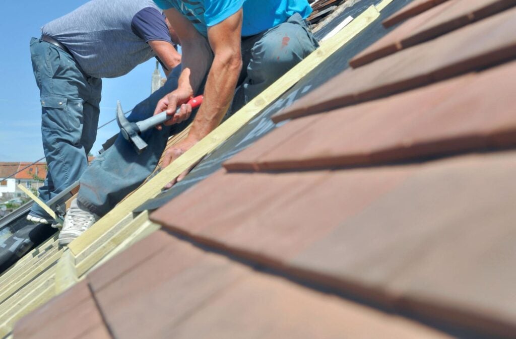 Two roofing contractors kneel on a sloped roof, installing insulation and tiles under a clear sky—ideal for Hook Agency's website.