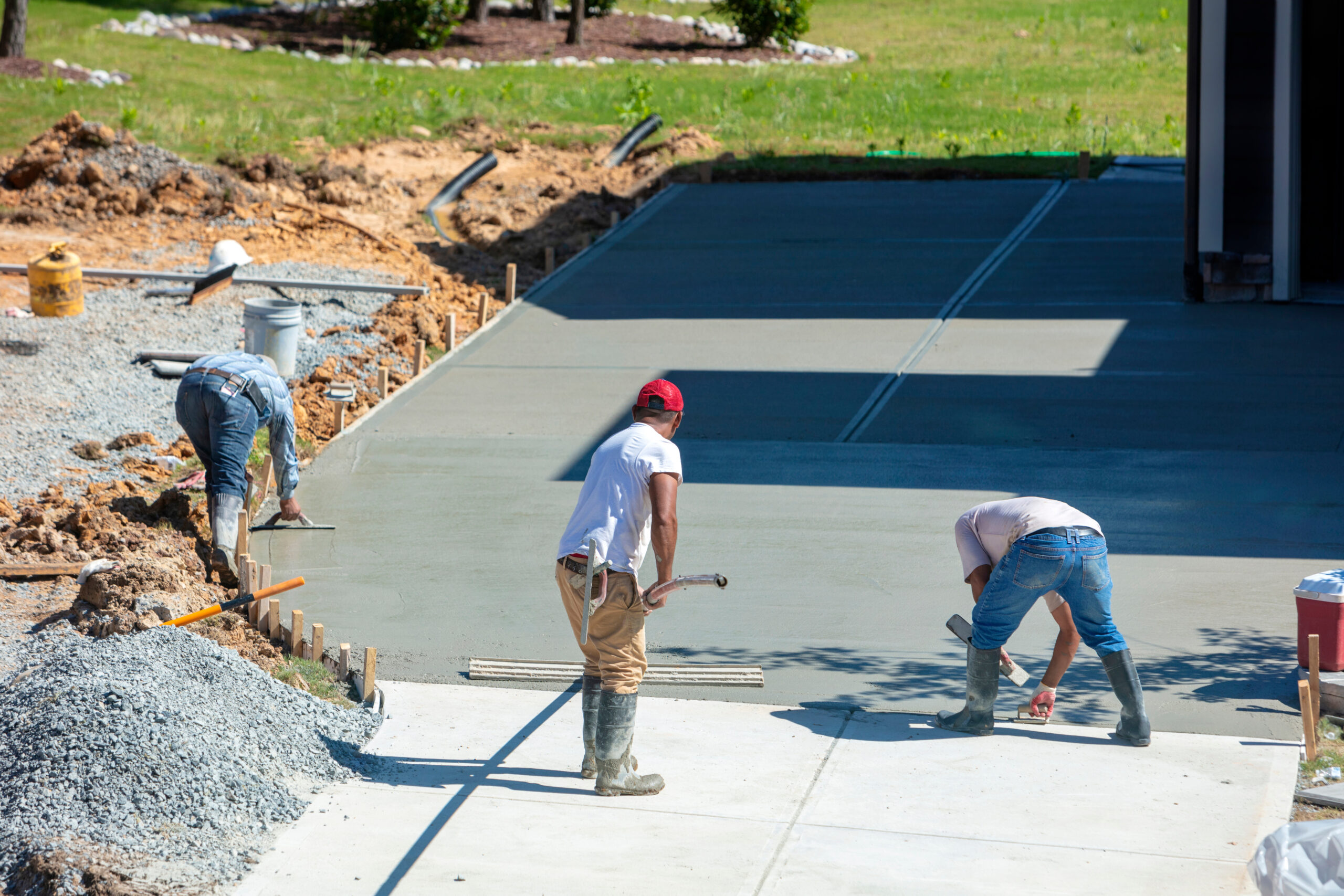 Three skilled construction workers expertly level and smooth freshly poured concrete on a driveway, embodying the art of hardscaping. Surrounded by gravel piles and tools, they work diligently in boots and casual attire, with a partially completed garage in the background.