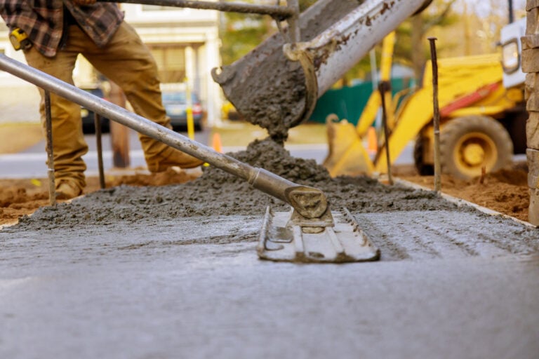 On Hook Agency's website: A construction worker expertly smooths wet concrete, crucial for the hardscaping project, while a yellow construction vehicle stands ready in the background.