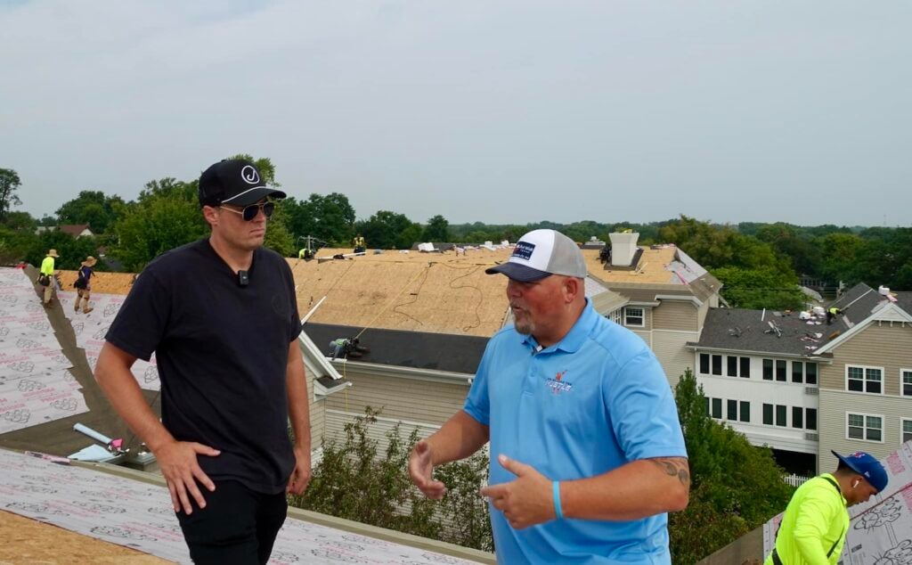 On a construction site, two men—one in a black shirt and sunglasses, the other in a blue polo and cap—stand on a roof discussing effective roofing sales strategies. Nearby, crews are actively working on neighboring rooftops.