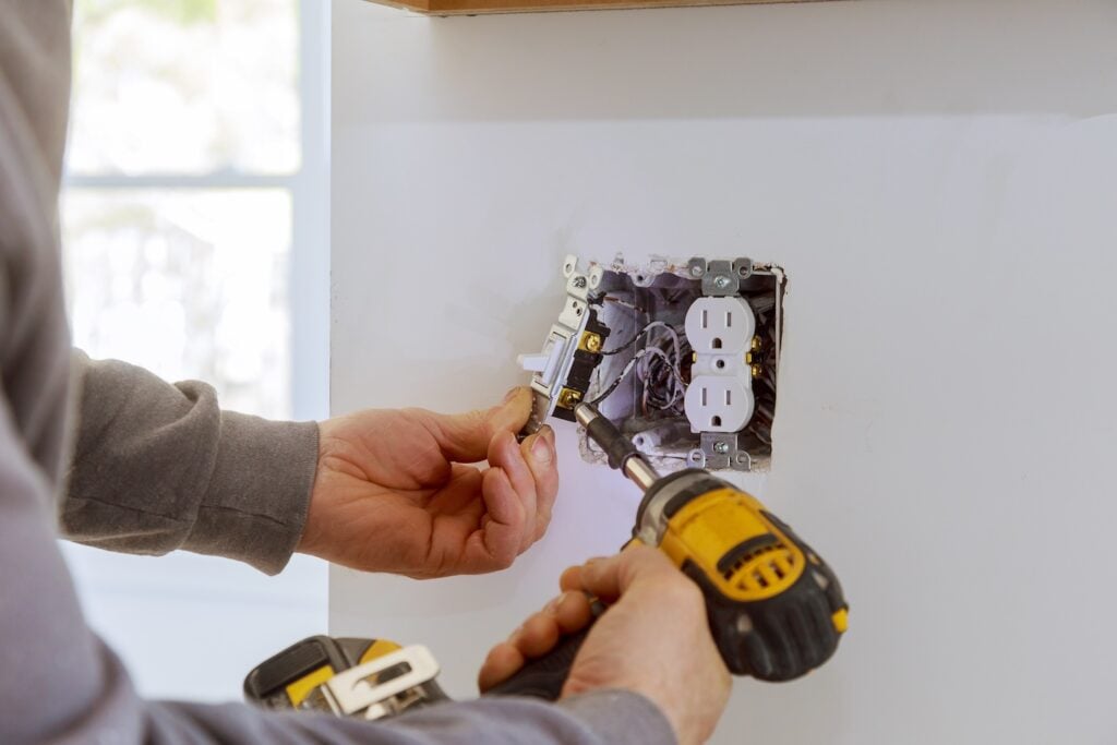 A technician uses a yellow cordless drill to install or repair a light switch beside a double electrical outlet on a white wall. Exposed wiring is visible within the electrical box, illustrating a standard home services task.