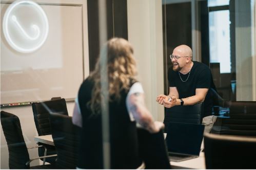 Two professionals engage in a dynamic discussion about contractor marketing in a modern, glass-walled meeting room. One stands, smiling with enthusiasm, while the other sits at the table with laptops open—collaborating on innovative strategies.
