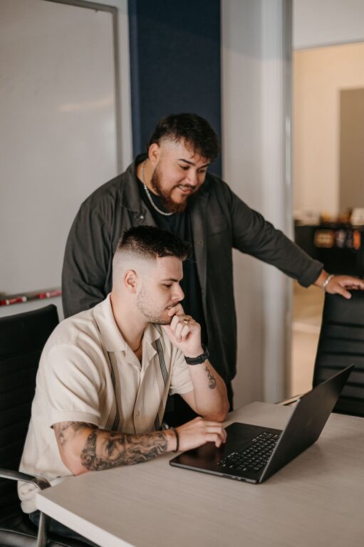 Two men in a professional office review a laptop together—one seated and typing, the other standing and engaged—actively collaborating on an effective foundation repair marketing strategy.