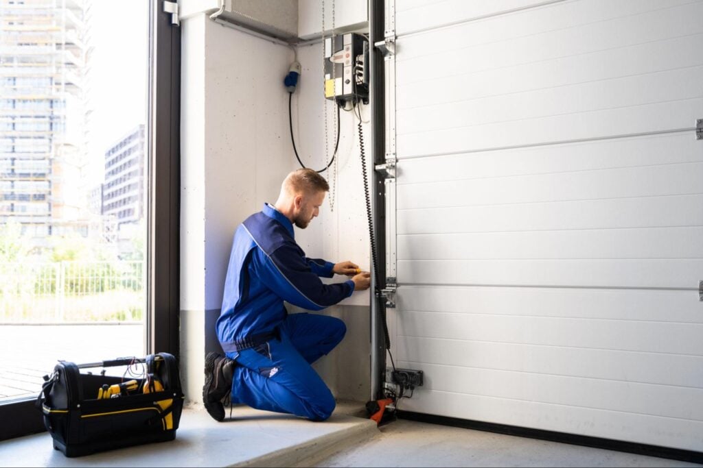 Expert technician in blue uniform repairs a white automatic garage door control unit; open black tool bag with yellow handles nearby.