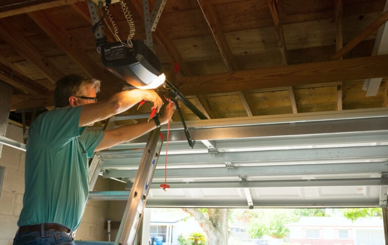 A person wearing a teal shirt stands atop a ladder, expertly repairing a garage door opener within a garage featuring modern floor coating. Sunlight streams through the partially open door, illuminating the wooden beams and open ceiling—a perfect setting to highlight cutting-edge garage website design. Visit Hook Agency’s website for innovative designs that transform ordinary spaces into extraordinary showcases.