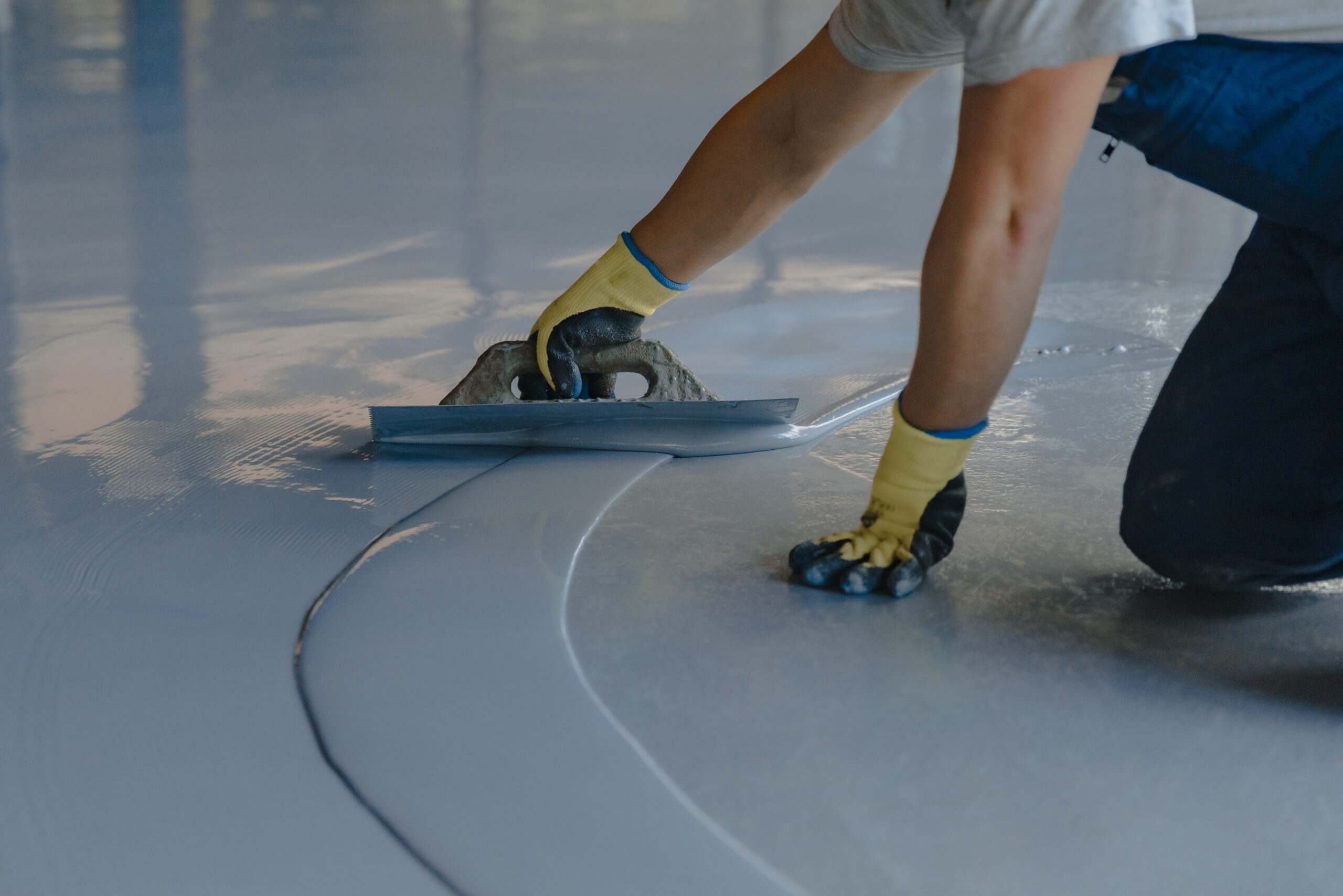 A person wearing yellow gloves uses a hand trowel to meticulously smooth gray epoxy garage floor coating, creating a reflective surface that is both functional and visually appealing.