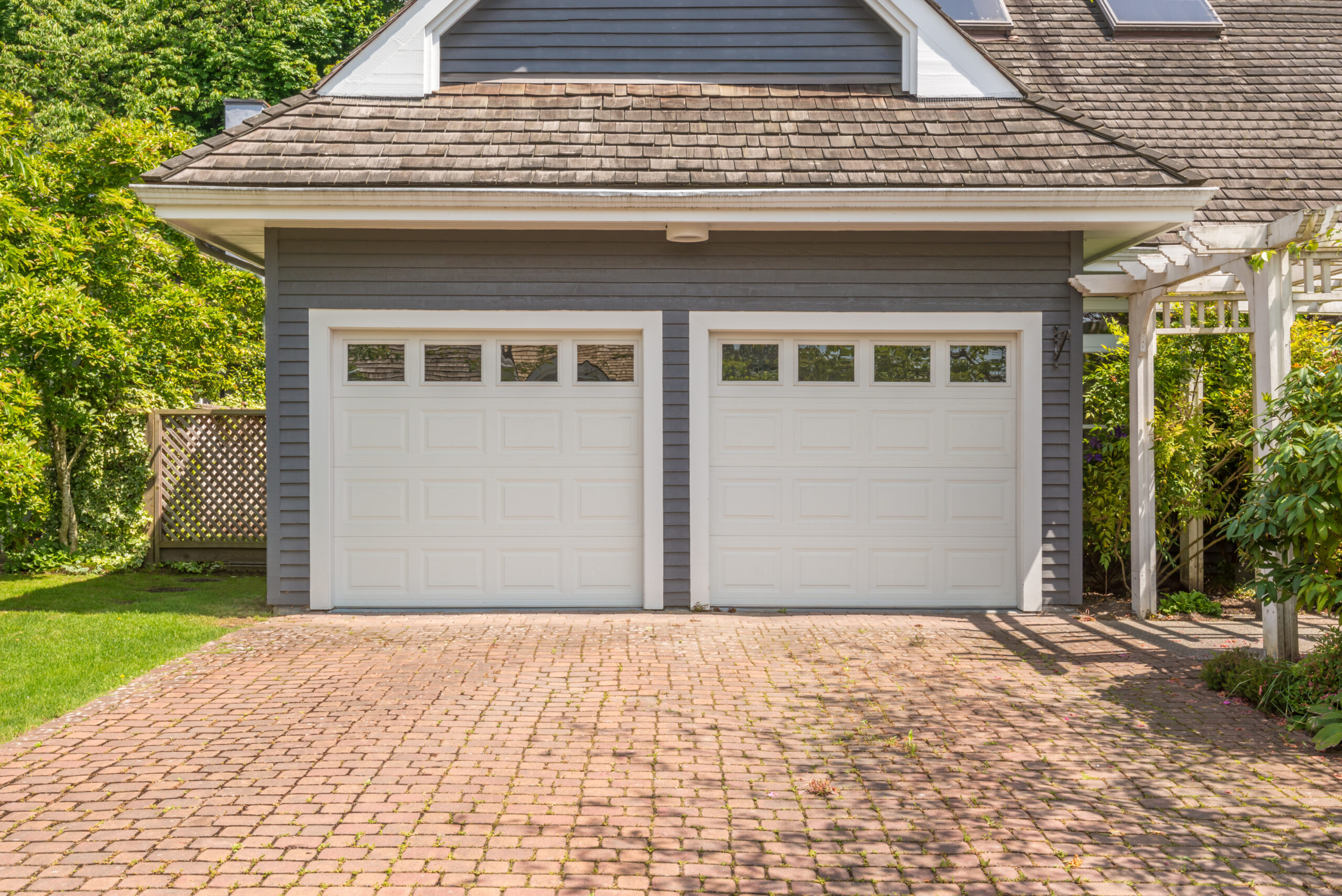 Showcasing superior garage construction, this two-car garage with closed white doors is seamlessly integrated into a gray house. The driveway, paved with red bricks and bordered by lush green bushes and trees, complements the design. Wooden shingles on the roof further elevate the home's aesthetic appeal.