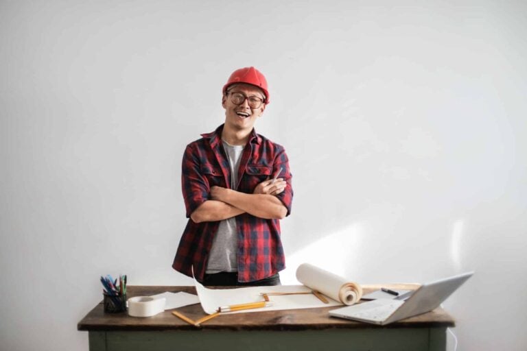 A contractor, equipped in a red hard hat and checkered shirt, stands confidently with arms crossed. Positioned before a desk filled with papers, a laptop, and drafting tools, he symbolizes readiness to optimize projects like Google Local Service Ads—all against a clean white backdrop.