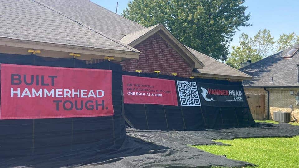 A house undergoing renovation prominently showcases three banners: one stating "BUILT HAMMERHEAD TOUGH," another featuring a QR code, and a third with a hammerhead logo. A black tarp partially conceals the structure, while trees frame the scene under a clear sky—illustrating an innovative approach to roofer marketing aimed at attracting roofing leads.