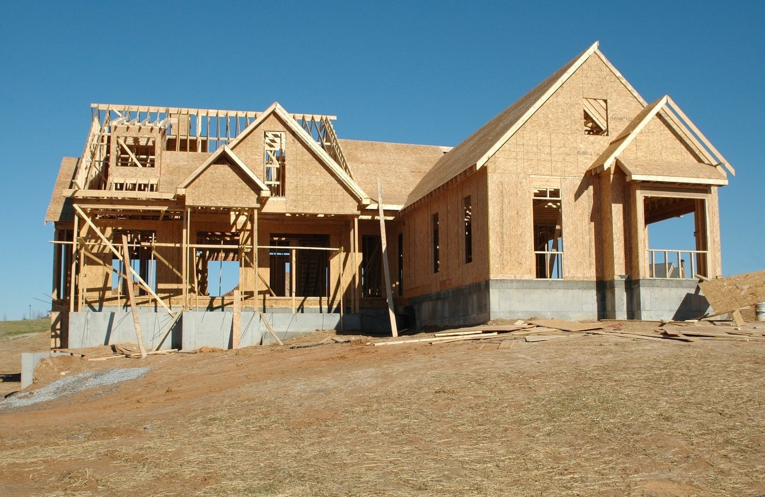 Under clear blue skies, a house under construction displays its wooden framework and partial roofing on a dirt lot. Scattered building materials underscore the expertise of the skilled contractors at work.