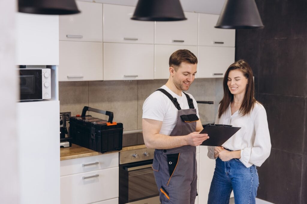 On Hook Agency's website, a technician in overalls discusses home service options with a woman in her kitchen. They stand amidst white cabinets and a countertop holding a microwave and toolbox, clipboard in hand. Both are smiling as they explore lead generation opportunities.