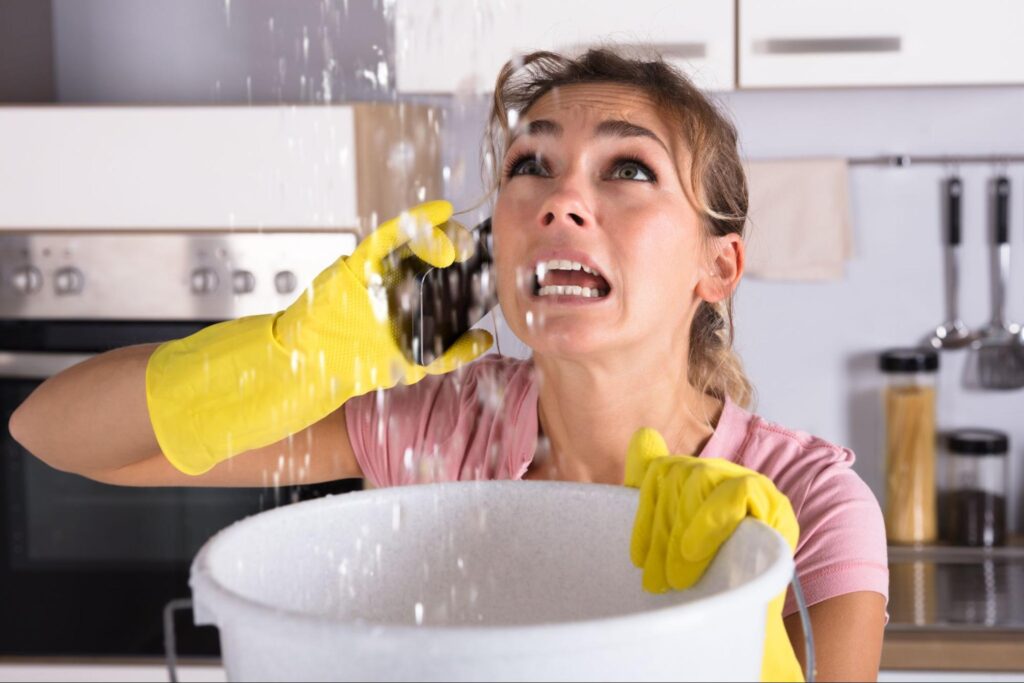 Woman in yellow gloves holds bucket under ceiling leak, phones for plumbing help in kitchen—address water damage immediately.