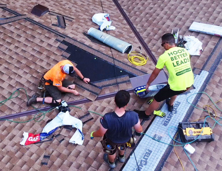 Three workers expertly install roofing shingles on a pitched roof, showcasing an impressive display of teamwork. One wears an orange shirt and cap, another dons a bright yellow shirt with text that says Hook Better Leads with HookAgency.com, and the third opts for gray. Tools and materials are scattered around, capturing the essence of roofing marketing at its best.