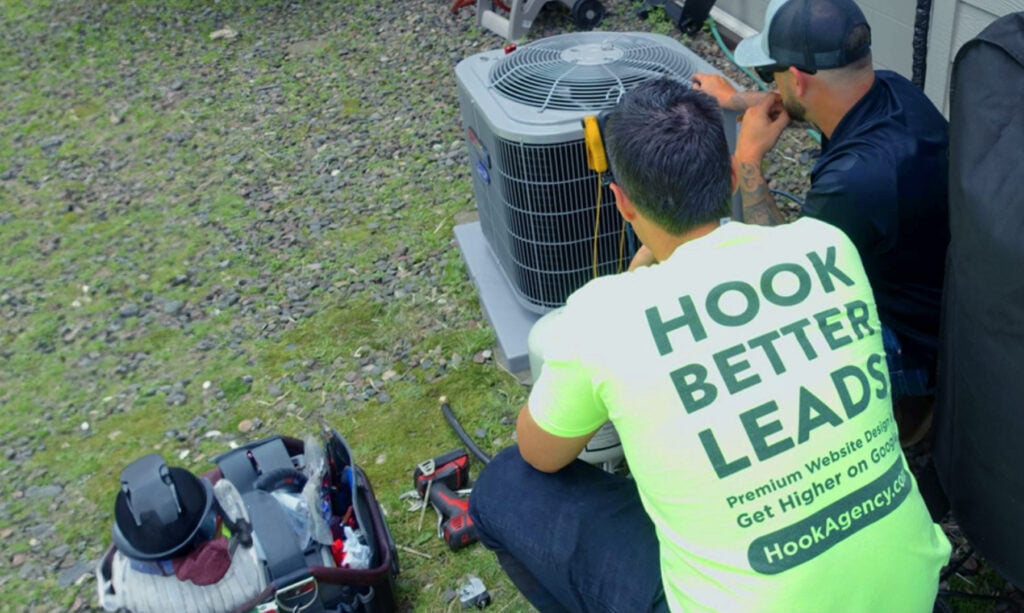 On Hook Agency's website: Two technicians concentrate on an outdoor HVAC unit, exemplifying successful HVAC marketing strategies. One technician sits on the gravel in a bright neon company shirt, as the other kneels beside him. Nearby, their tool bag and equipment are strategically placed, enhancing the professional setup.