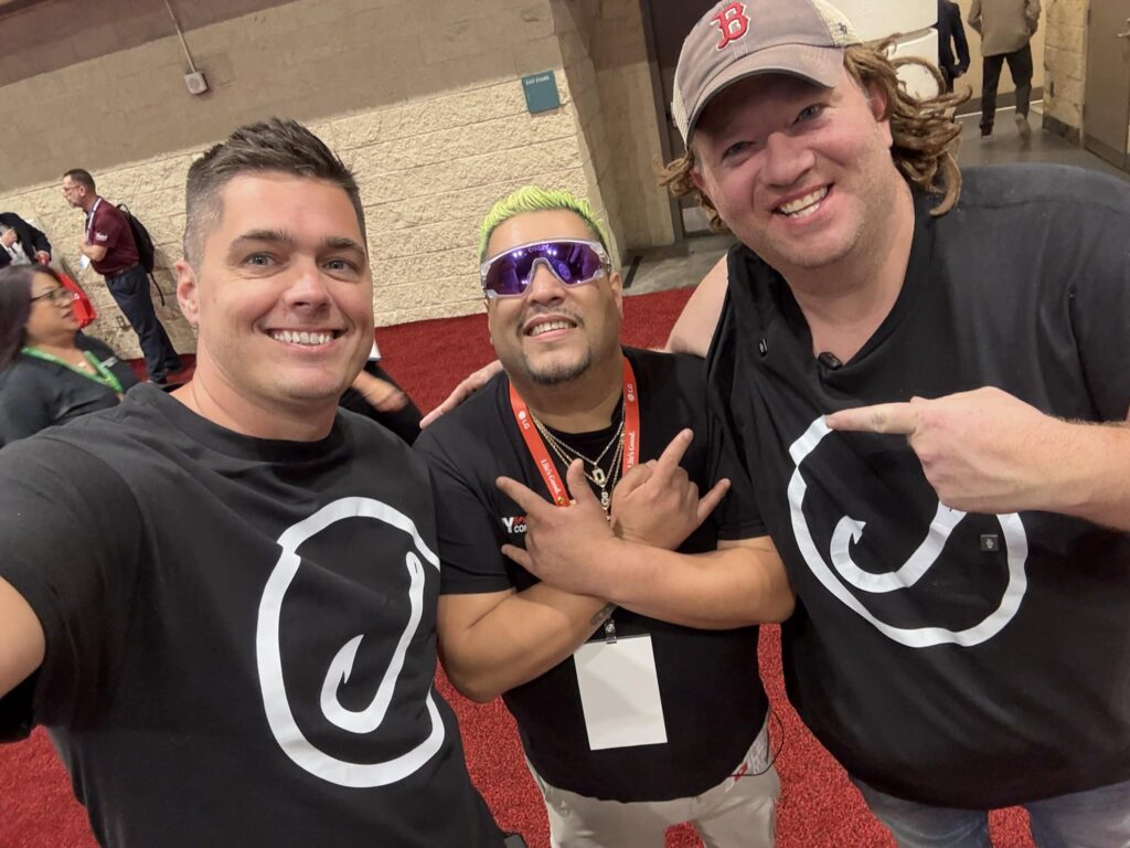 Three men pose for a selfie on a red carpet at an indoor event. Two HVAC professionals wear matching black shirts featuring the white Hook Agency logo; one wears a Boston Red Sox cap. The third man, sporting blond hair and sunglasses, flashes hand signs. Other attendees network in the background.