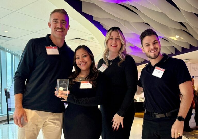Four business professionals pose indoors, one holding an award, under purple-lit modern ceiling—celebrating career achievement.