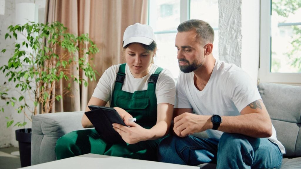 Technician in green overalls shows a tablet to a homeowner, highlighting HVAC mistakes to avoid, in a bright living room.