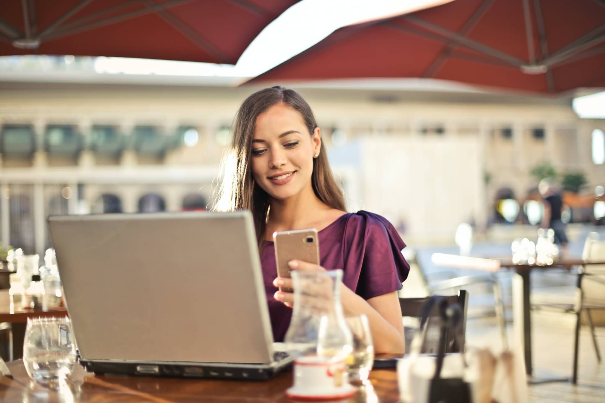 HVAC Schema Markup SEO A woman in a purple blouse sits at an outdoor café, smartphone in hand and laptop open. With a confident smile, she strategizes her next competitive SEO move under sunlit umbrellas.