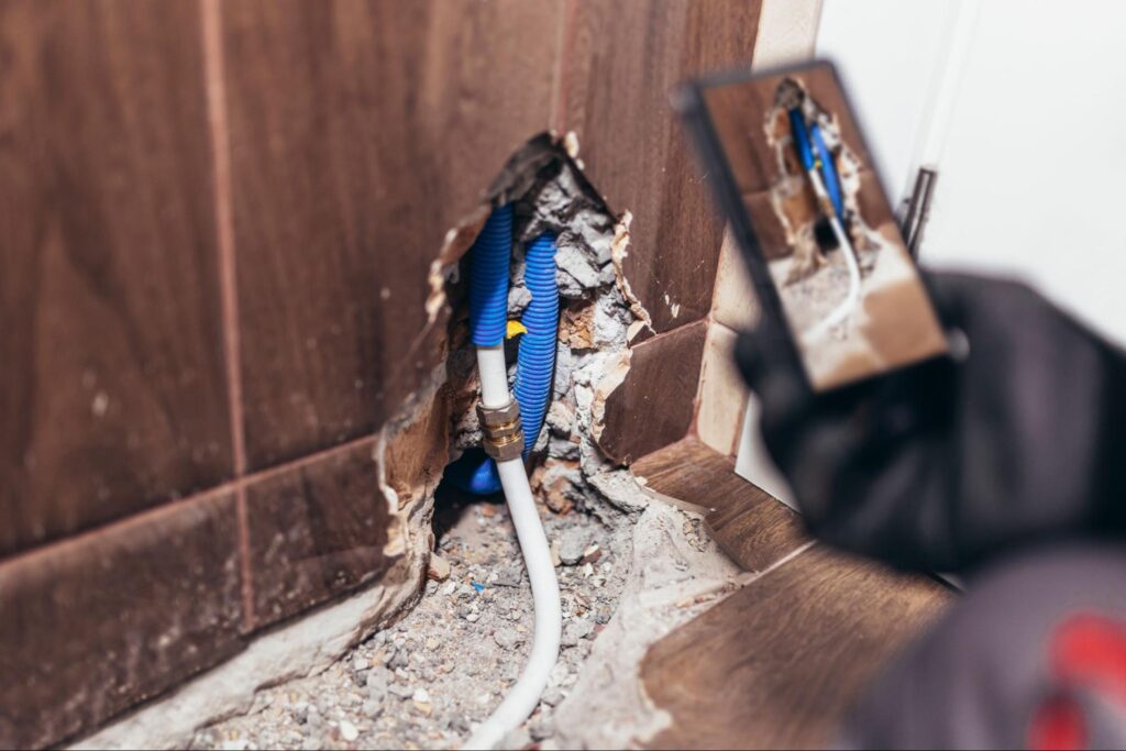 Person photographing a damaged wall with exposed pipes and debris, capturing the urgent need for professional plumbing repair.