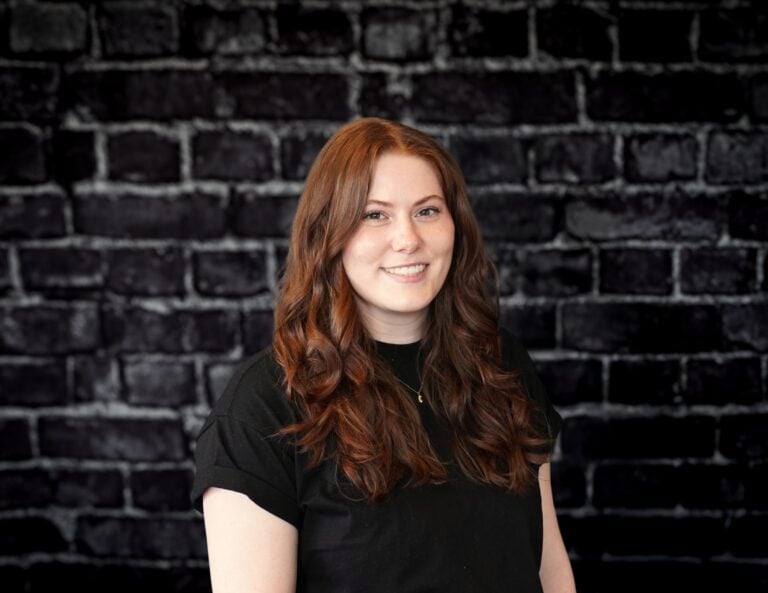 Madelynn Wald, team member at Hook Agency, smiles in front of a dark brick wall, wearing a black shirt and wavy hair.