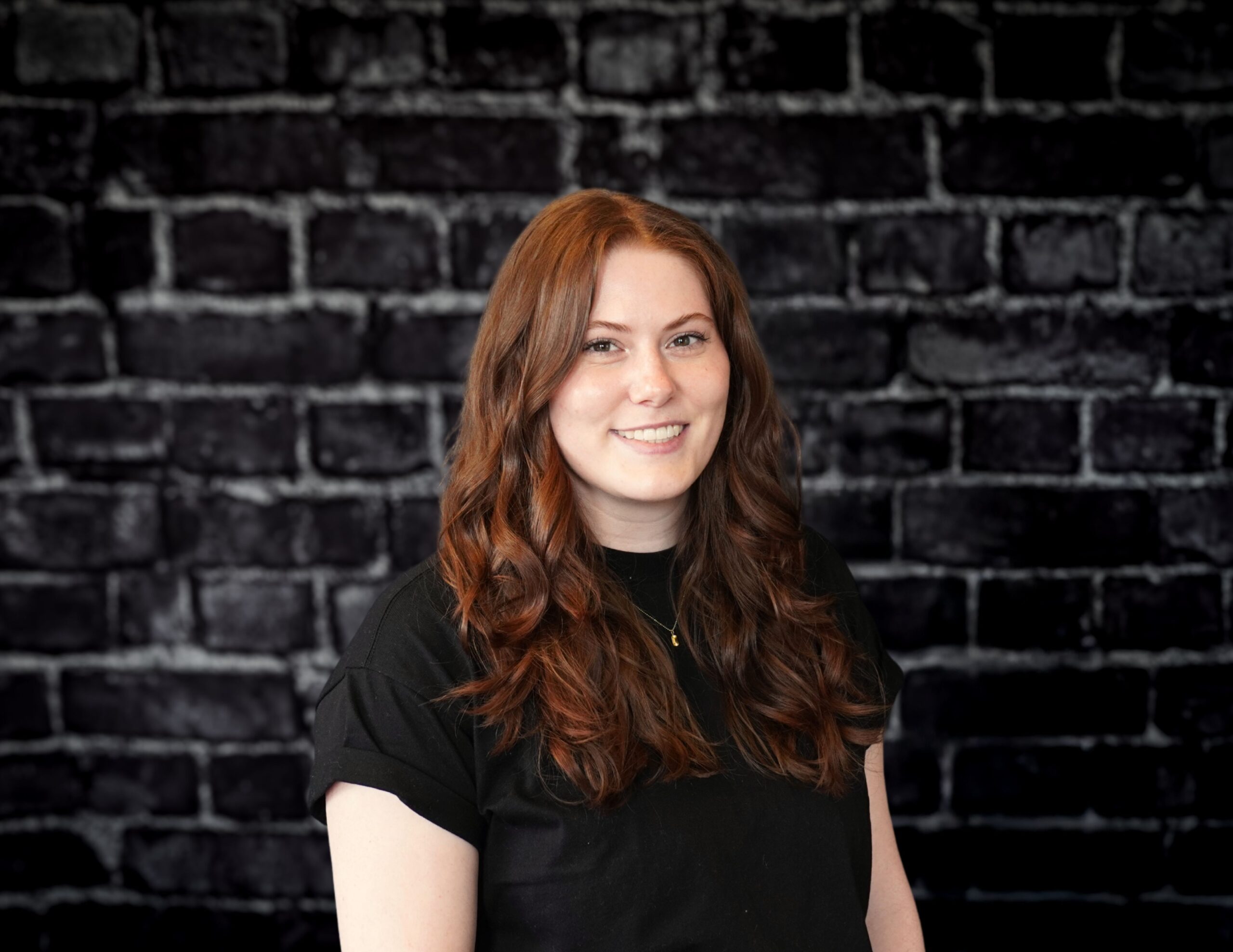 Madelynn Wald, team member at Hook Agency, smiles in front of a dark brick wall, wearing a black shirt and wavy hair.