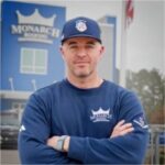 Martin Pettigrew stands confidently with arms crossed outside the Monarch Building store, wearing a branded blue Monarch sweatshirt and cap. The Monarch Building logo and his name are clearly displayed on the storefront behind him.