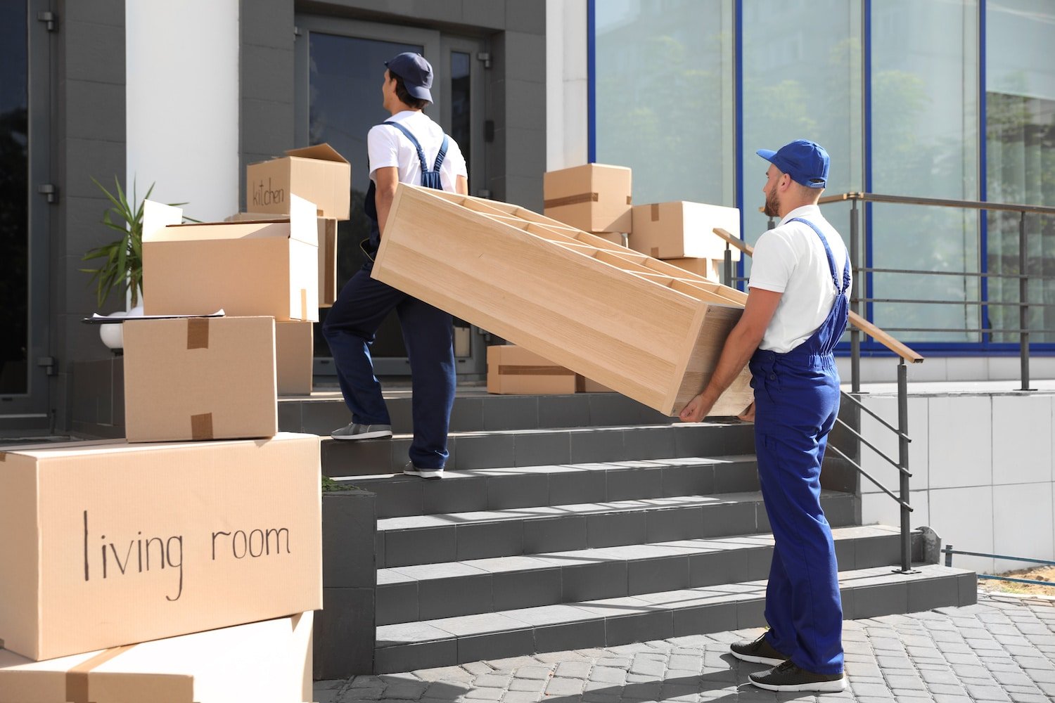 Two movers, dressed in blue uniforms, efficiently carry a wooden shelf up the steps into a house. Labeled cardboard boxes, including one for the "living room," are arranged strategically around them. This scene illustrates moving day at a modern building entrance, highlighting our professional moving company's unmatched efficiency and expertise.