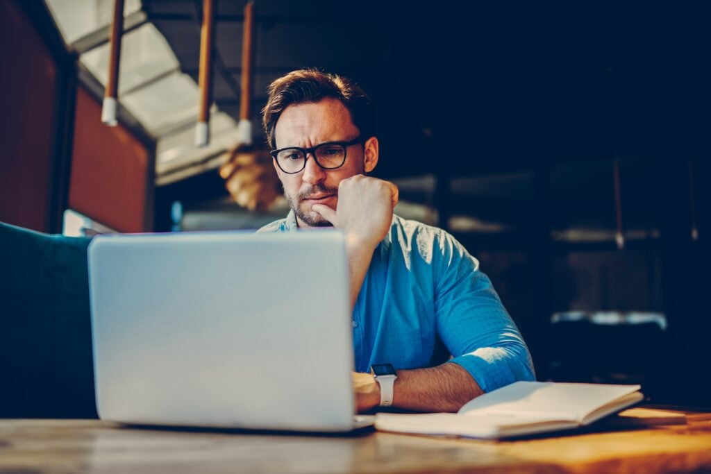 A man in glasses and a blue shirt reviews SEO strategies at his laptop, thoughtfully considering next steps. An open notebook and pen are ready beside him for actionable insights.