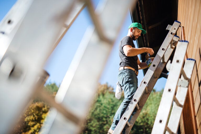 A man in a green cap stands on a ladder painting the exterior of a wooden building, captured from below against a clear blue sky. This image is ideal for enhancing your painting blog's content strategy.