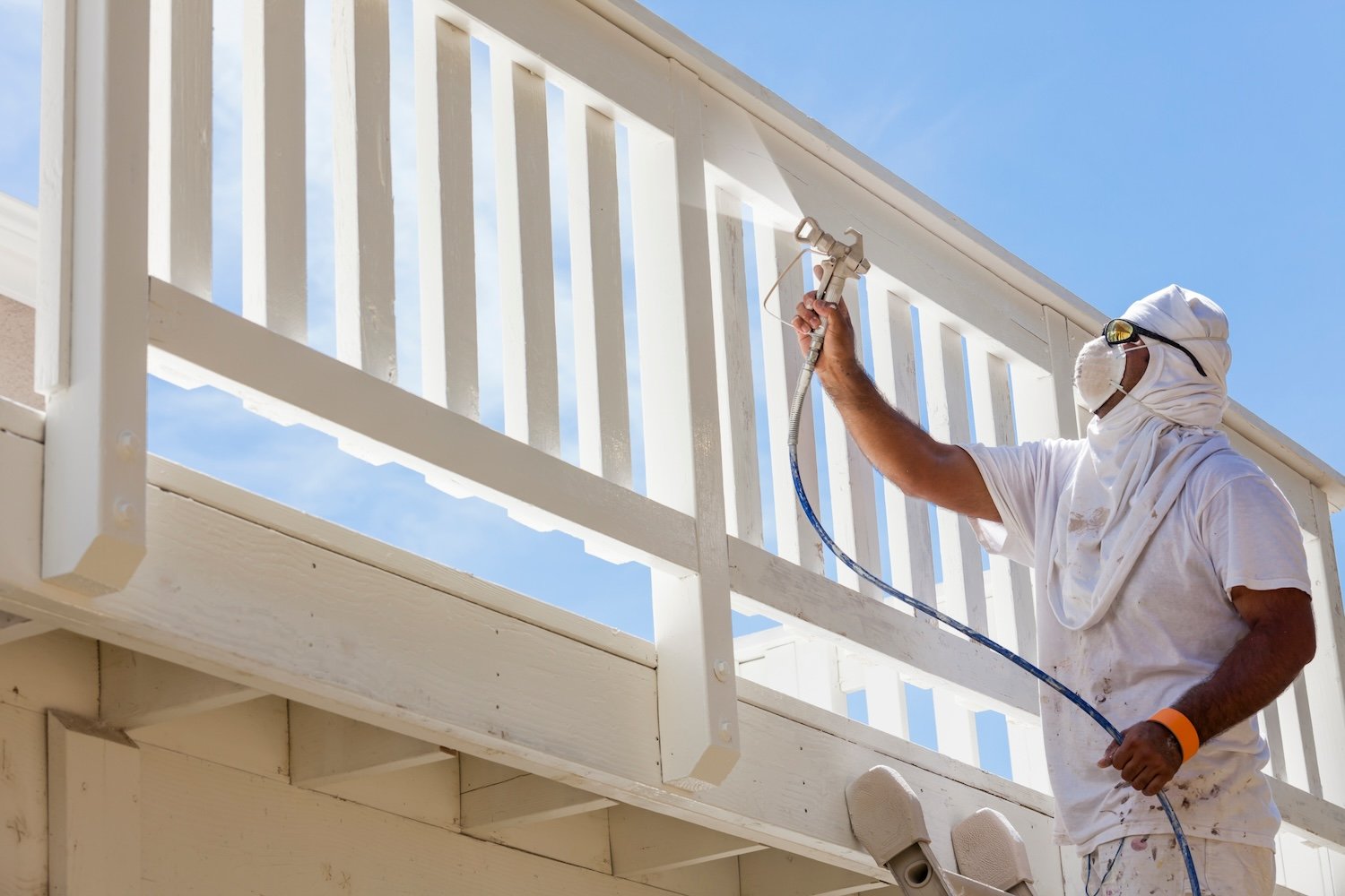 A person, equipped with protective clothing, mask, and goggles, is efficiently spray painting a white fence under a clear blue sky.