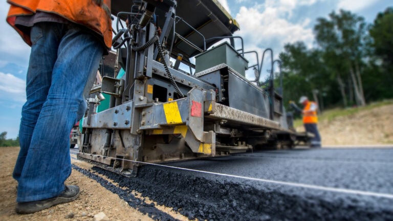 Witness the precision of an efficient paving operation as a road paving machine expertly lays fresh asphalt. Skilled workers, donned in safety gear, manage the equipment with diligence. A worker's legs are in focus, set against a backdrop of softly blending trees beneath a clear blue sky.