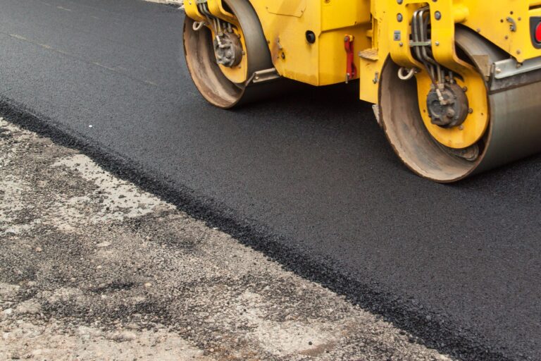 A yellow asphalt roller efficiently compacts a newly paved road, contrasting the rough gravel on the left with the smooth finish on the right.