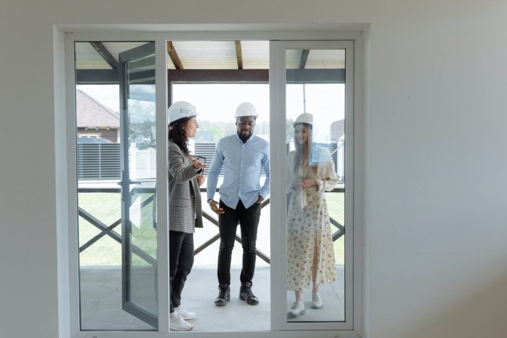 Three individuals in hard hats stand on a porch, engaged in a discussion about roofing solutions. Positioned in front of glass sliding doors, two women and a man are strategizing lead generation approaches for their businesses, with houses and lush greenery in the background.