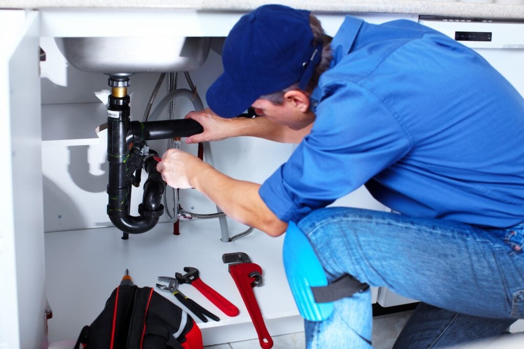 A professional plumber in a blue uniform and cap works beneath a kitchen sink, using a wrench to repair black piping—a classic example of profitable home service work. Essential tools like wrenches and pliers are organized on the floor, ready for efficient use.
