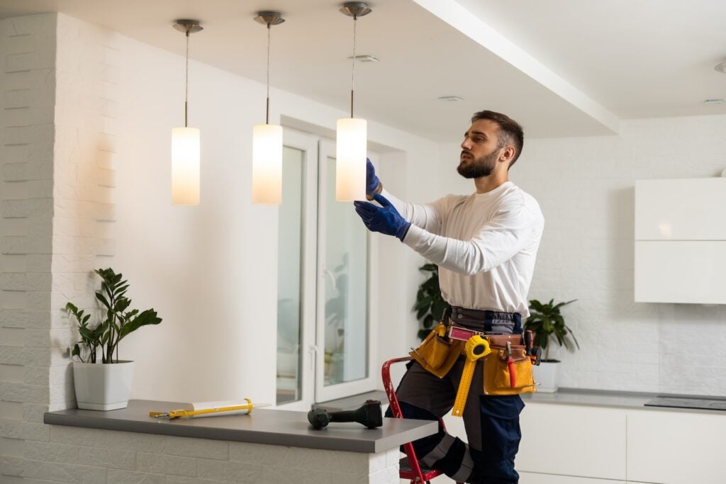 A licensed electrician in safety gear stands on a step stool, expertly adjusting a kitchen light fixture. Surrounding tools and plants emphasize the range of specialized electrical skills required for precise installations like this.