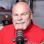 Roger Wakefield, a well-known industry expert, sits confidently in front of a microphone. He is easily recognized by his bald head, prominent white mustache, and signature red shirt. The background features blurred shelves and a brown cap, highlighting the professional yet approachable atmosphere.