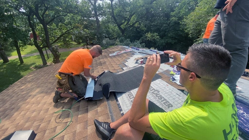 Roofing Leads - Two roofers on a shingled roof are actively engaged in their craft. While one expertly installs shingles, the other documents the process with a smartphone to enhance roofing SEO. Clad in casual work attire, they operate under a clear sky with trees providing a natural backdrop. Best roofing marketing companies