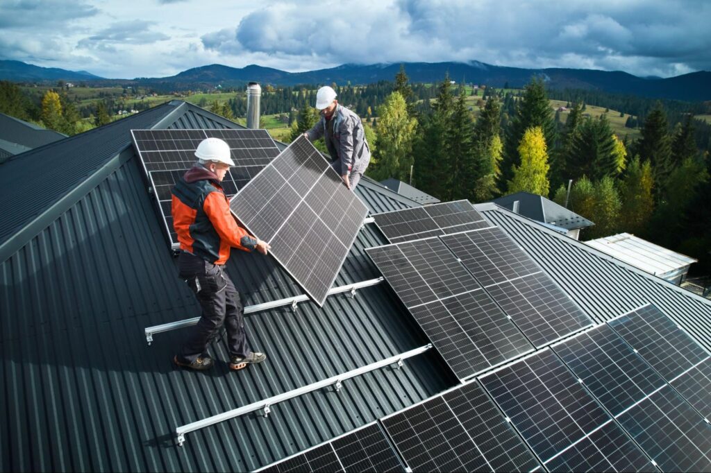 Workers in safety gear install solar panels on a sloped metal roof, set against forested mountains and a cloudy sky.