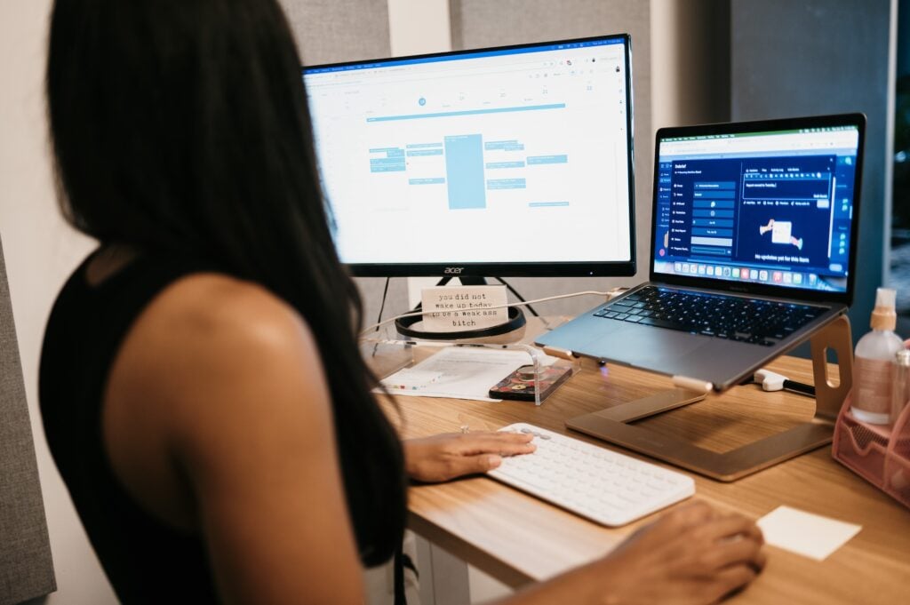 Person at desk reviewing SEO progress on large monitor and laptop; calendar, charts, office supplies, and sanitizer visible.