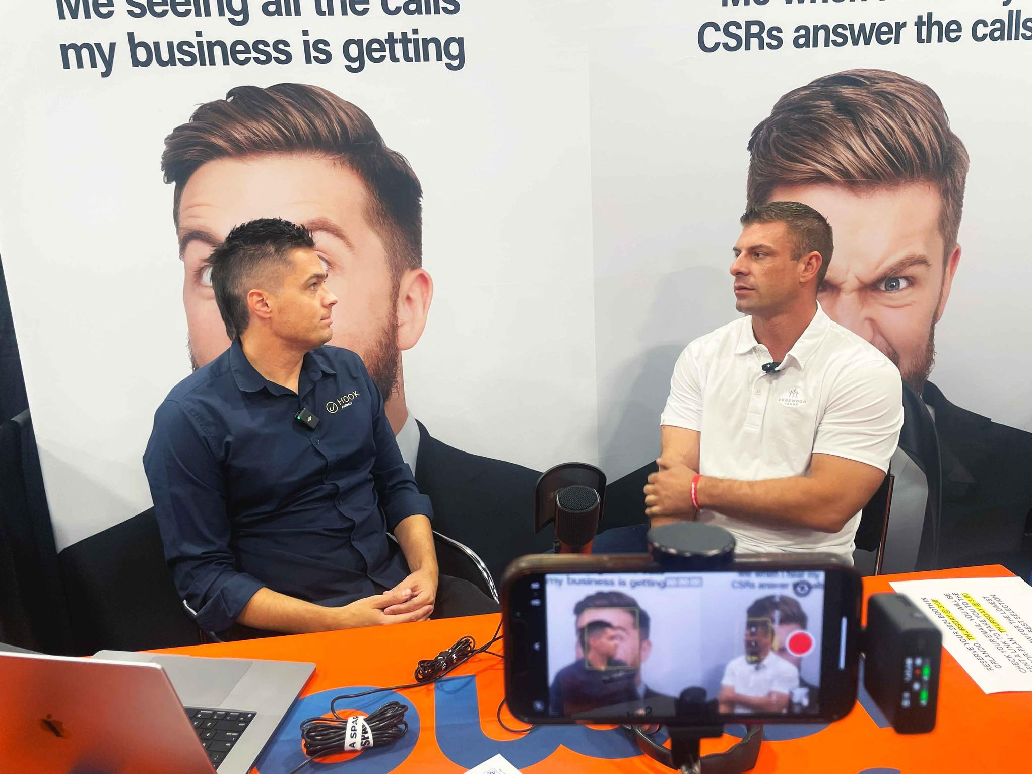 Two men sit at a table engaging in a discussion captured on video. One wears a dark shirt Tim Brown owner of Hook Agency, the other a white polo, Tommy Mello of A1 Garage Door. Behind them, a poster features a man with slicked-back hair and headset, subtly suggesting Garage Door SEO strategies. Nearby are a laptop, phone, and recording equipment.