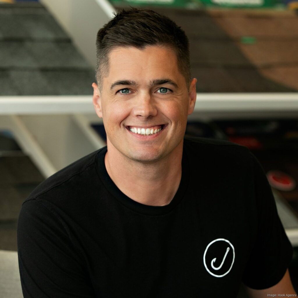 A man with short dark hair, dressed in a black shirt featuring a white circular logo, sits indoors surrounded by shelves. He contemplates what it takes to be the smartest person in Minnesota.
