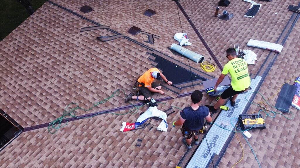 Three workers install shingles on a sloped brown roof, surrounded by tools and supplies—ideal imagery for Hook Agency marketing.