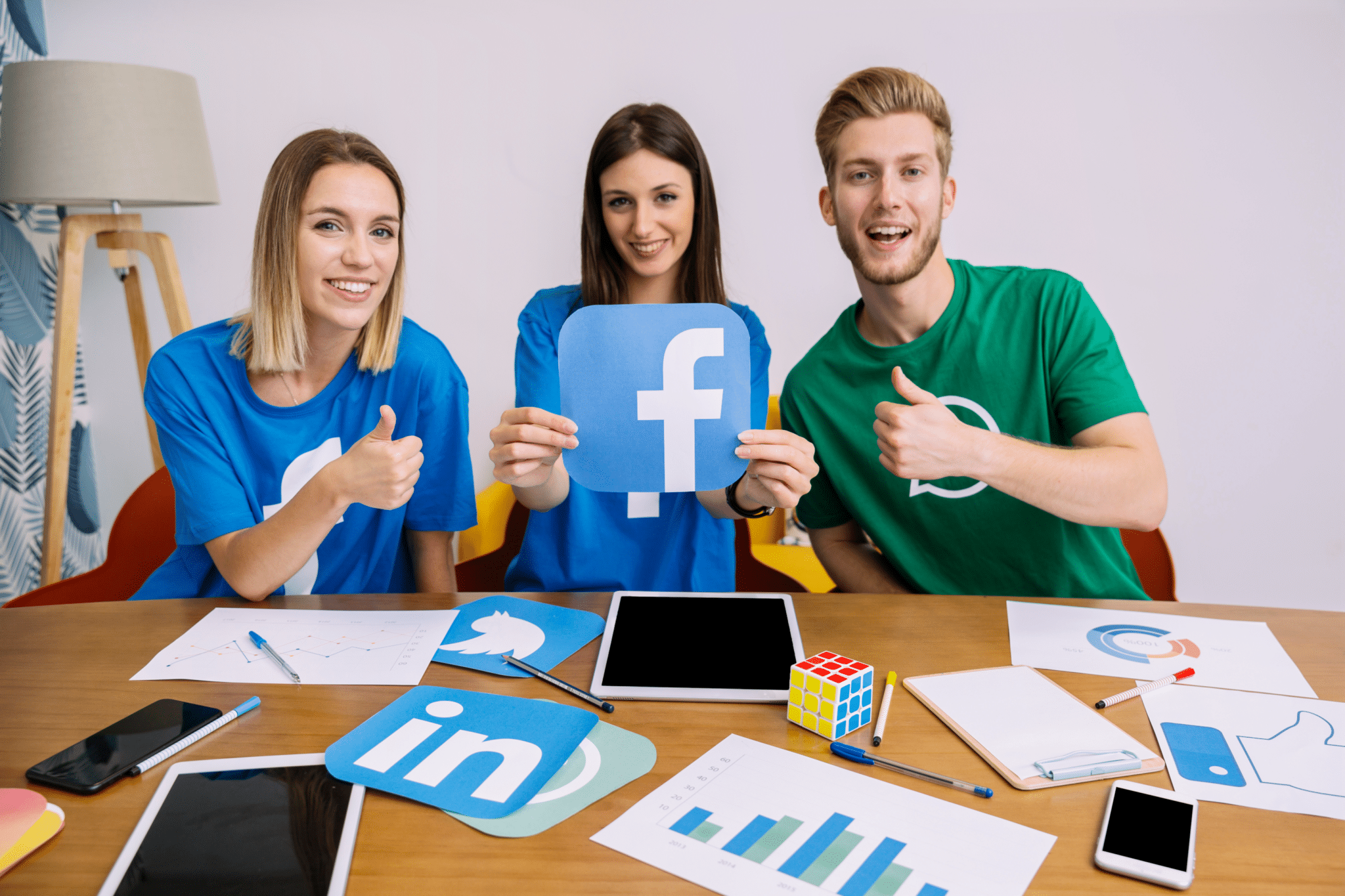 One woman holding the Facebook logo, while her 2 friends, 1 woman and 1 man are holding their thumbs up.